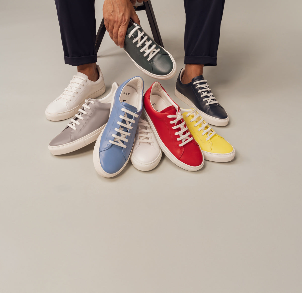 A hand lifting a green sneaker from a pile of red, yellow, blue, grey, white and navy JAK sneakers