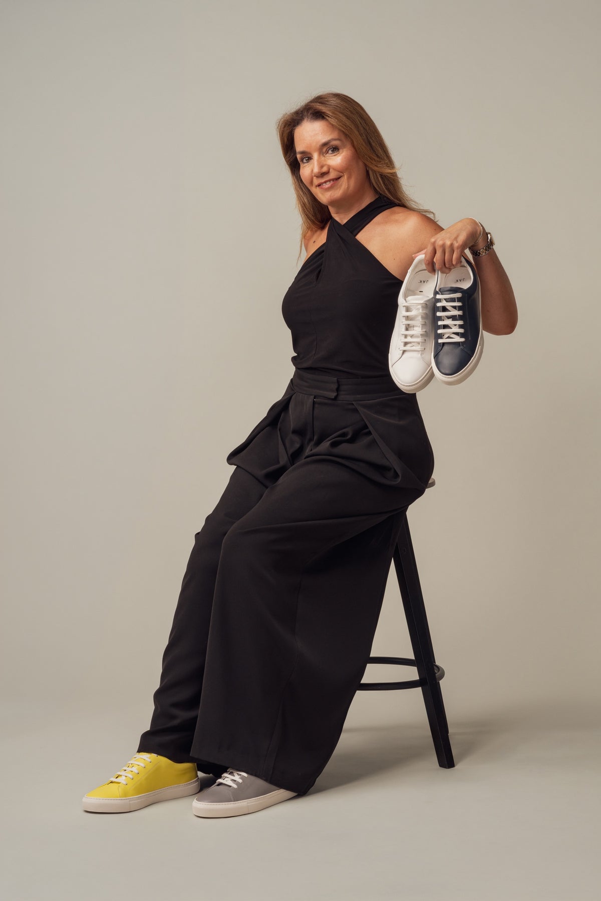 A woman in black on a stool holding up a sneaker, wearing one yellow and one grey shoe